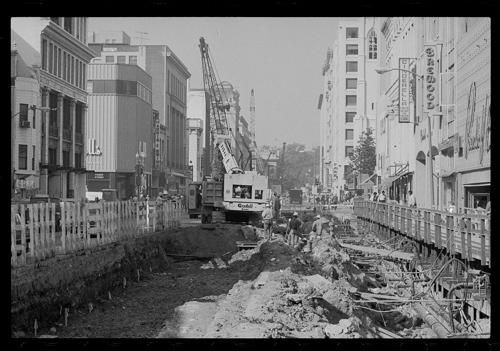 A black-and-white photo of Metro construction. There is a hole in the middle of a street with buildings on either side. There are construction workers and vehicles in and around the hole.