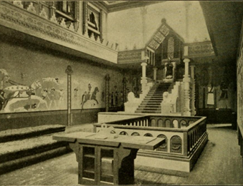A photo of the Assyrian throne room in the Hall of the Ancients. In view is a large throne and canopy with a model seated in it. Over the walls are paintings