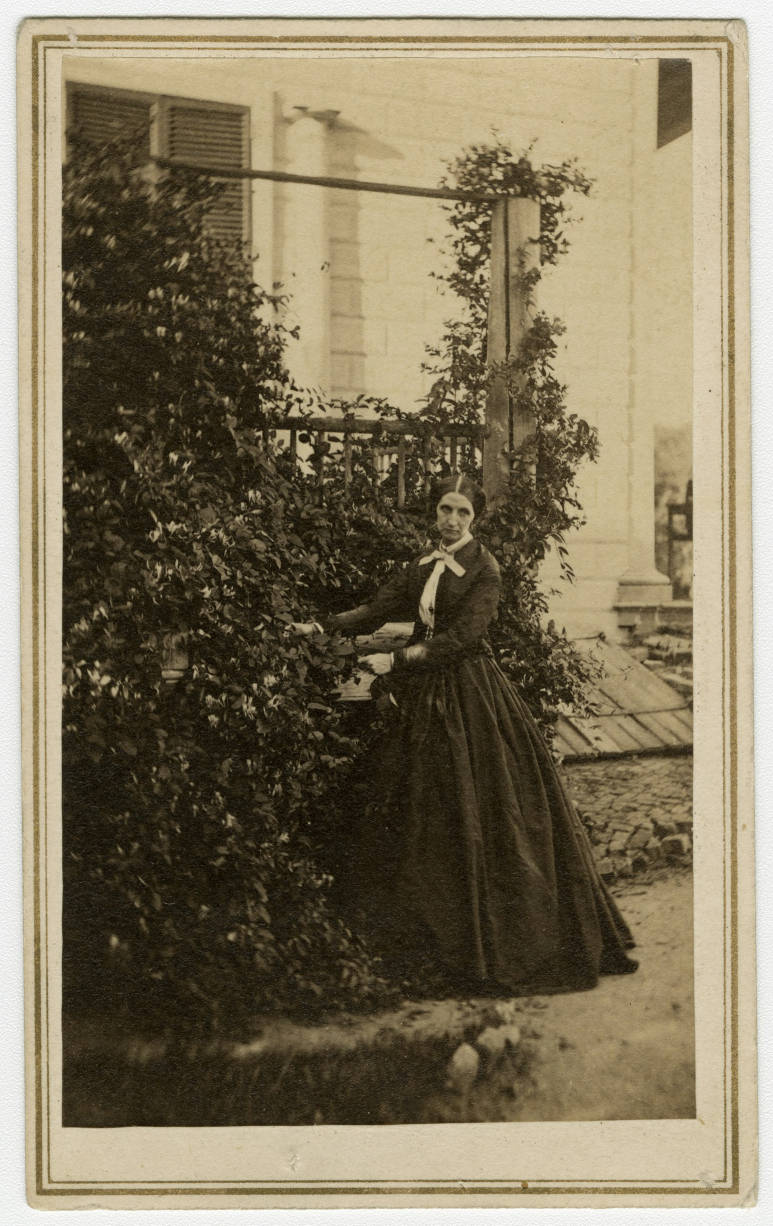 A photo of Sarah Tracy standing under a climbing plant in front of one of Mount Vernon's outbuildings