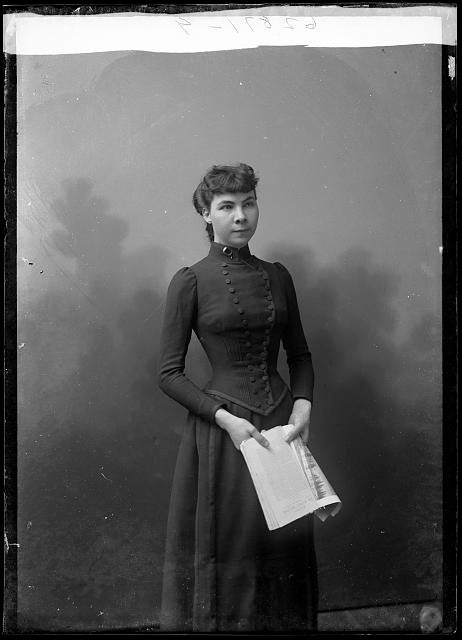 A 3/4 length photo of Madeline Pollard standing in a black dress with a newspaper in her hands