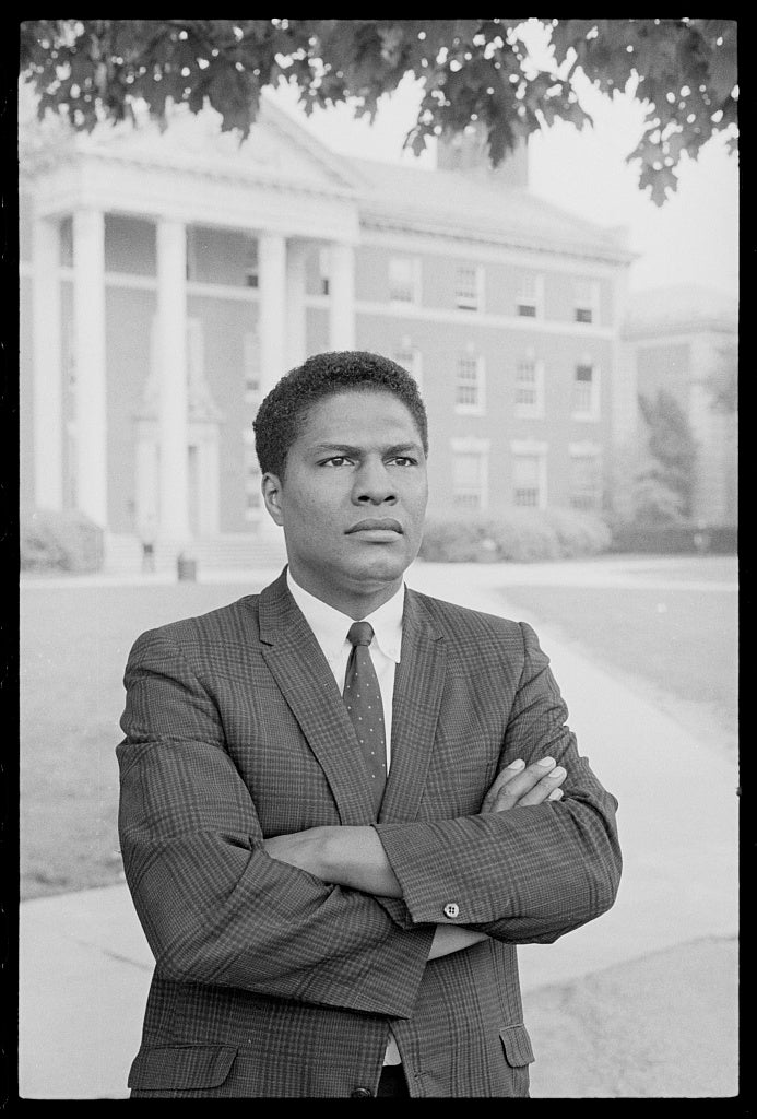 Professor Nathan Hare posing in front of Howard University's Douglass Hall dressed in suit with arms crossed.