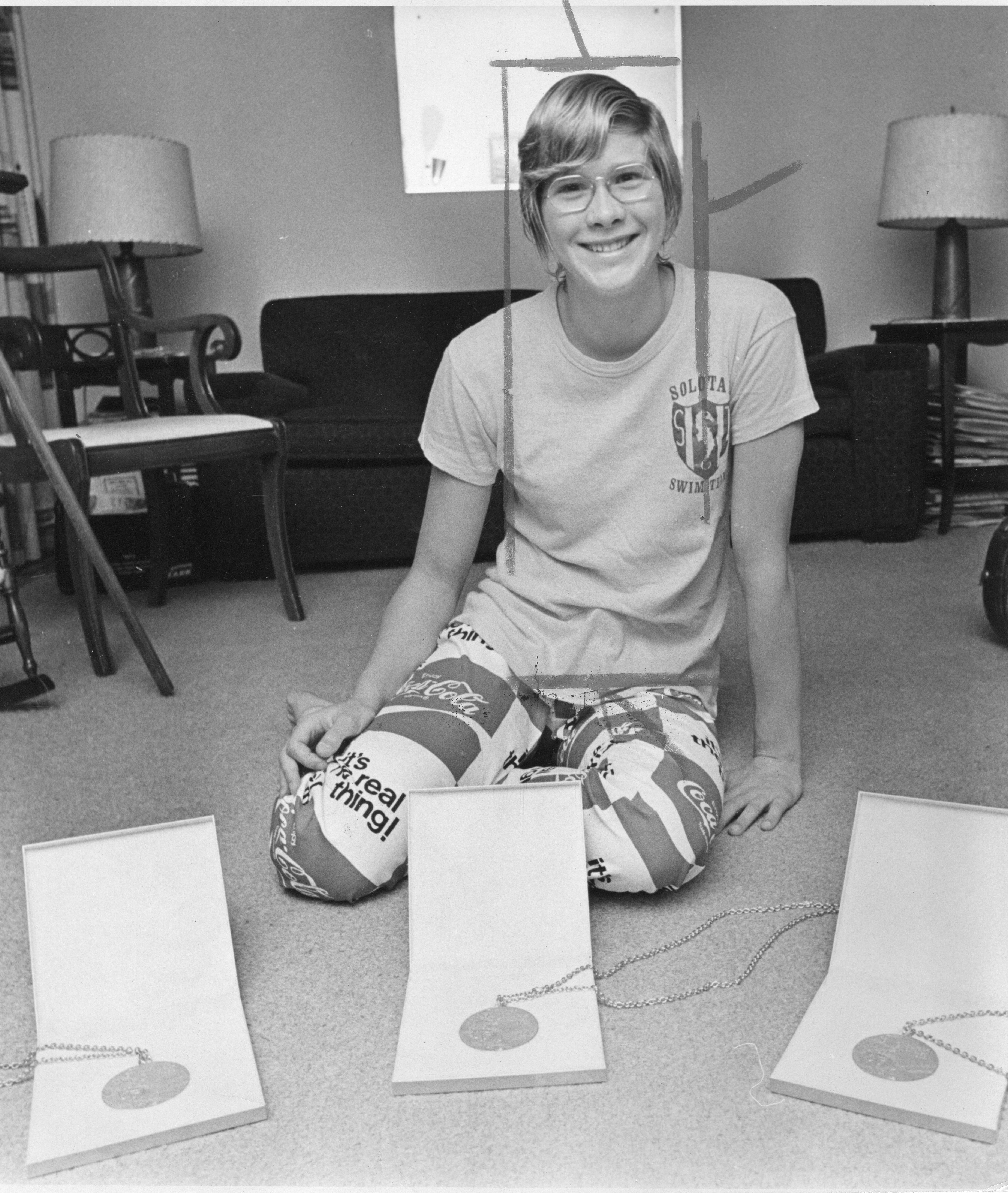 Melissa Belote sits on the floor at home with three gold medals on the floor in front of her.