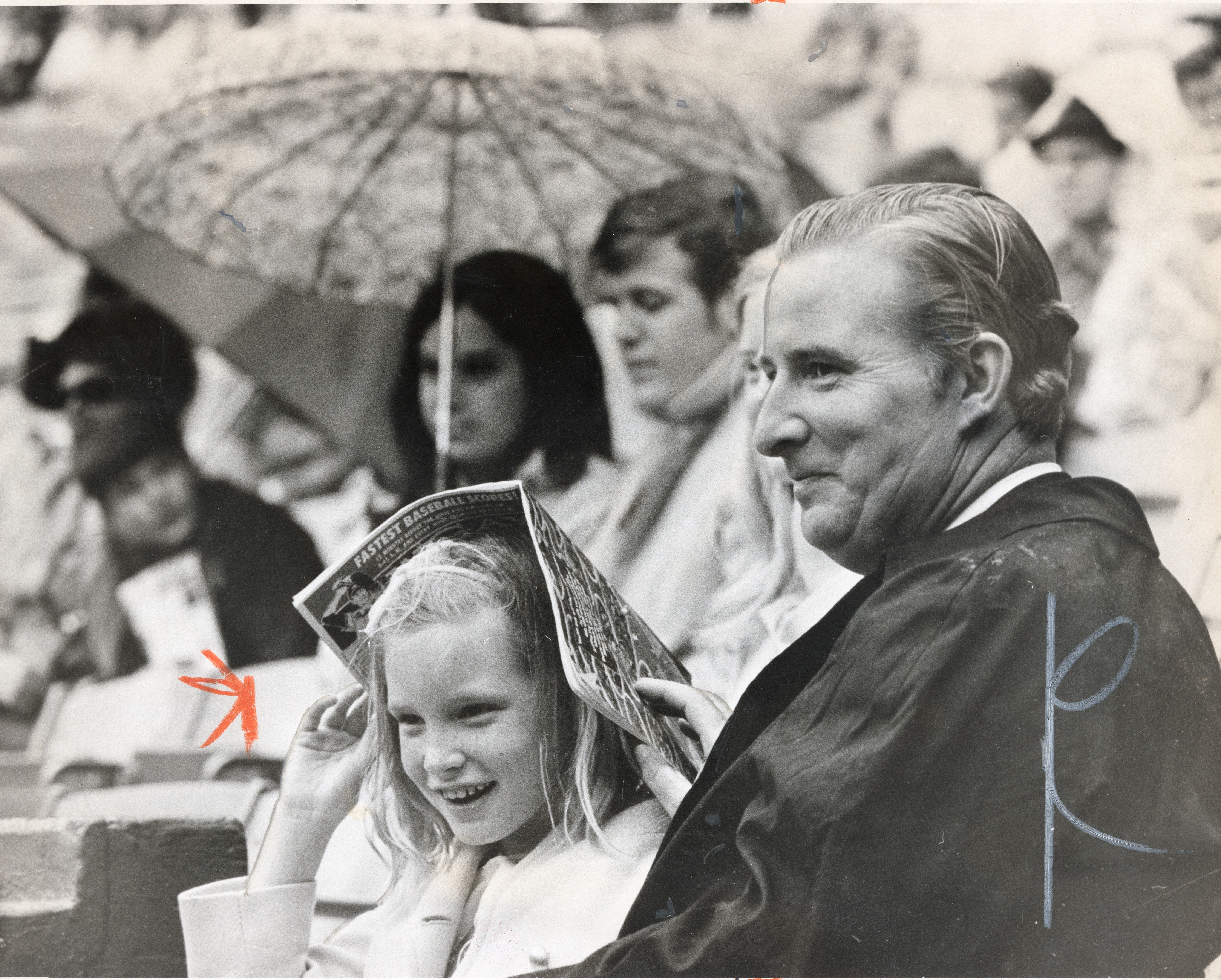 A young girl sits in the rain, covering her head with an open magazine. To her right, her dad sits smiling.