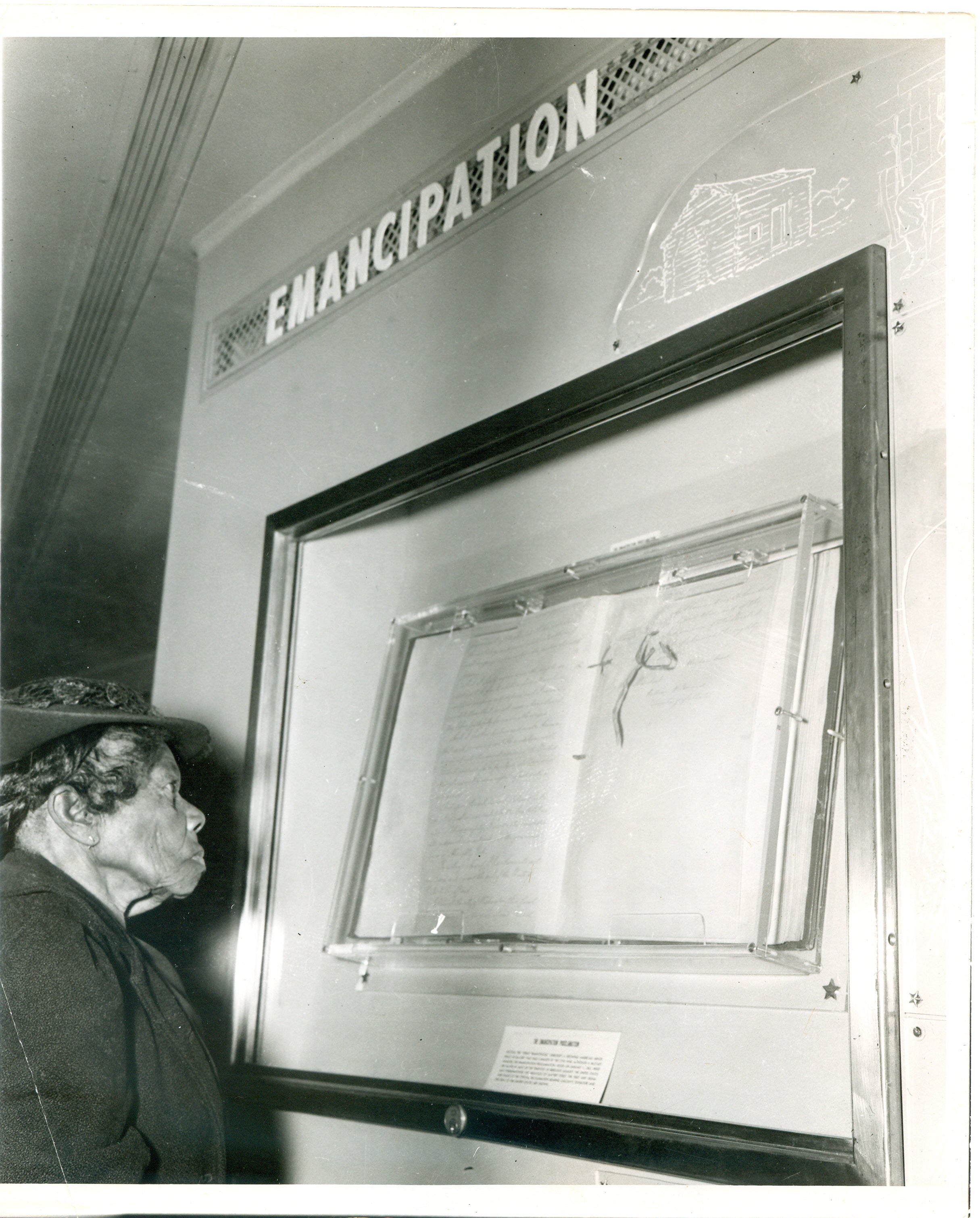 Elderly African American woman look at document in case on the wall. The word Emancipation is displayed over the case.