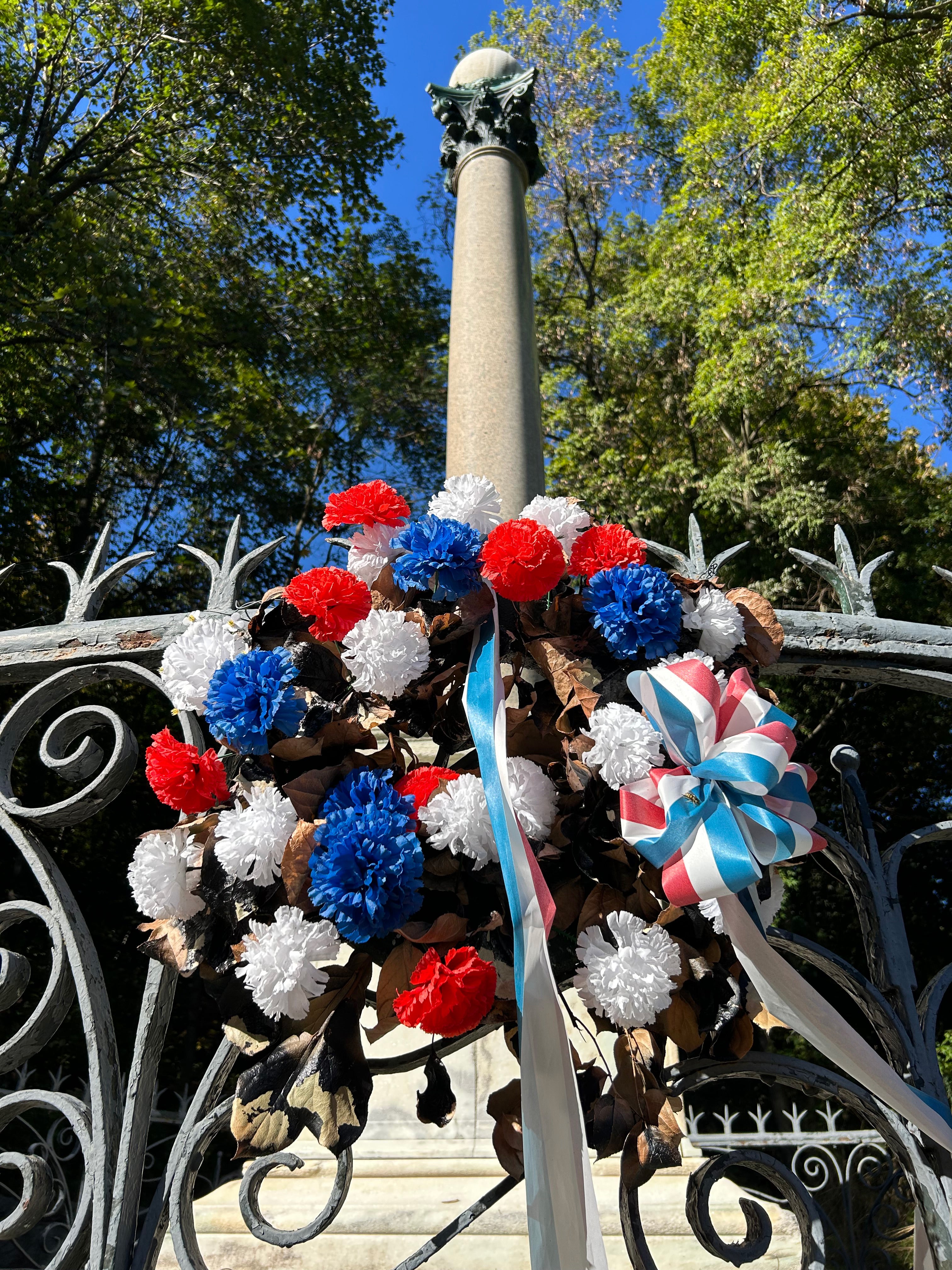 Tall cylindrical monument surrounded by wrought iron fence adorned with red, white and blue flowers.