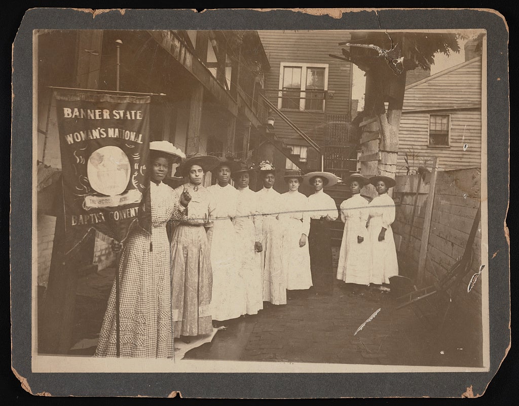 Nine African-American women posed, standing, full length, with Nannie Burroughs holding banner reading, "Banner State Woman's National Baptist Convention"