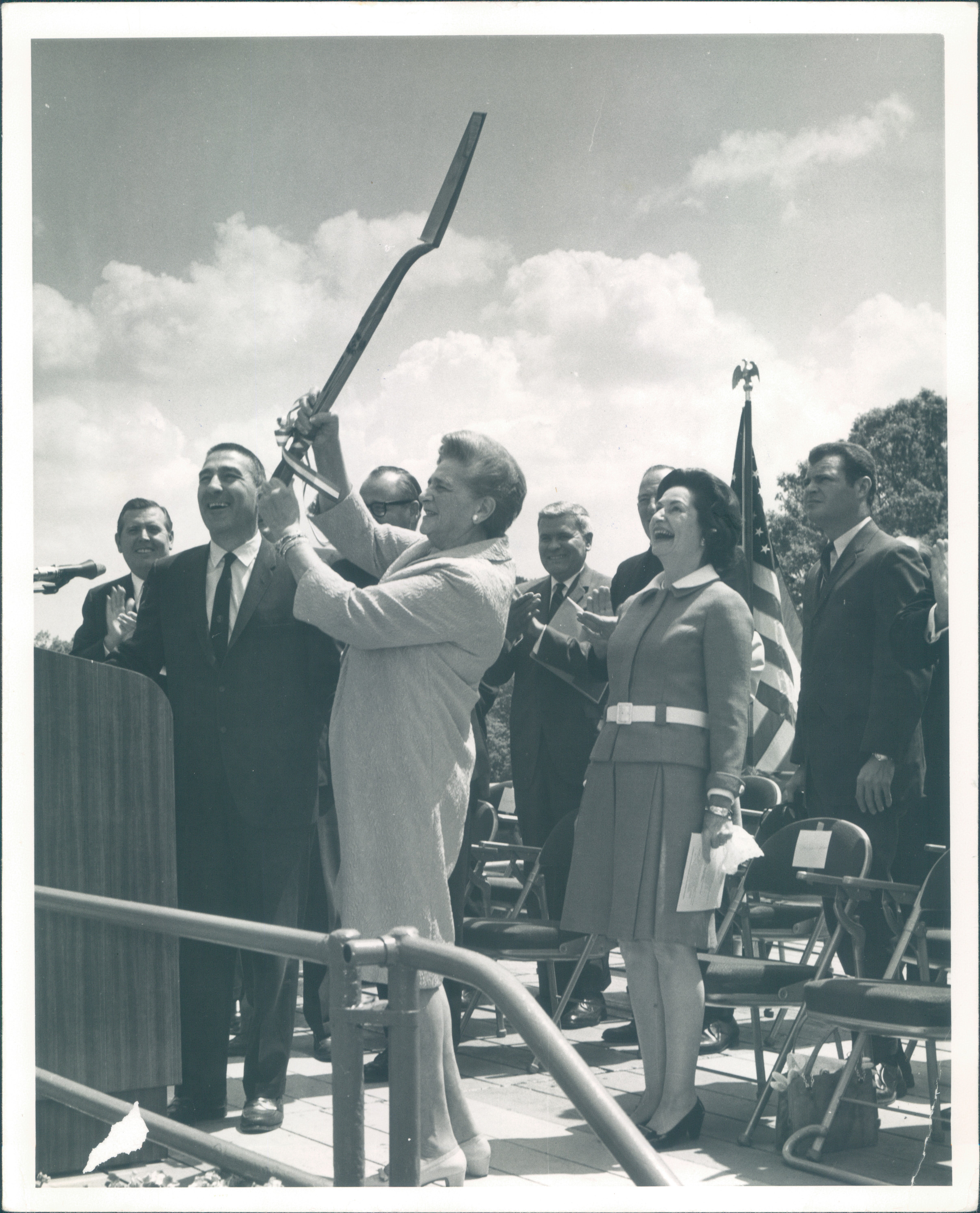 Woman stands on stage and holds shovel above her head as crowd behind her laughs.