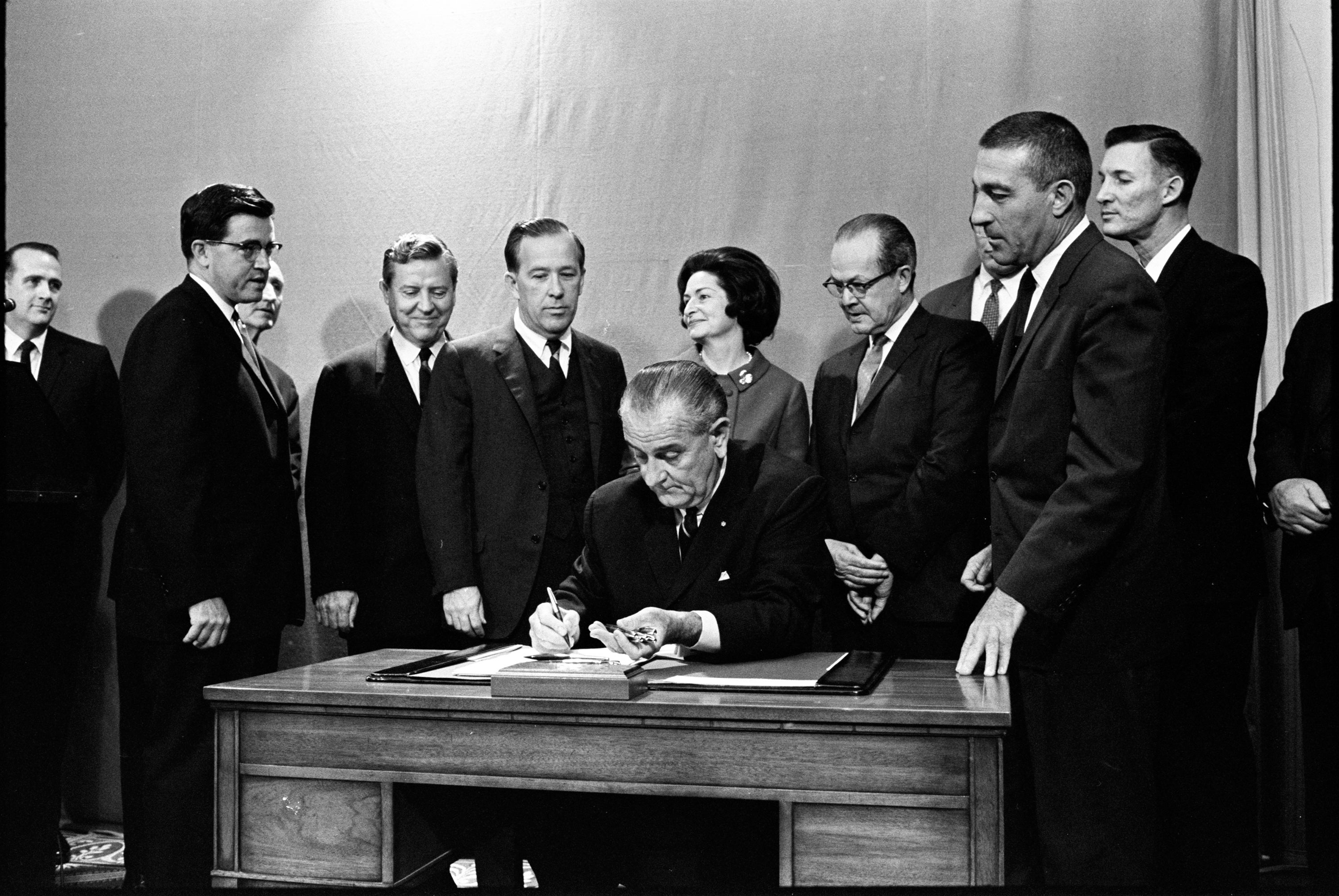 President Lyndon Johnson signs bill as a group of onlookers encircle his desk.
