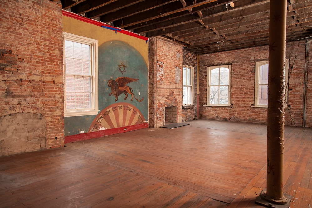 The mural is adjacent to a part of the floor that was removed to create a women’s balcony when 415 M was a synagogue. Although the floor boards were subsequently replaced, the visible incongruity indicates the outline of the earlier hole between the first and second floors. Patricia Fisher, Fisher Photography, 2014. The mural is adjacent to a part of the floor that was removed to create a women’s balcony when 415 M was a synagogue. Although the floor boards were subsequently replaced, the visible incongruity indicates the outline of the earlier hole between the first and second floors. Patricia Fisher, Fisher Photography, 2014.