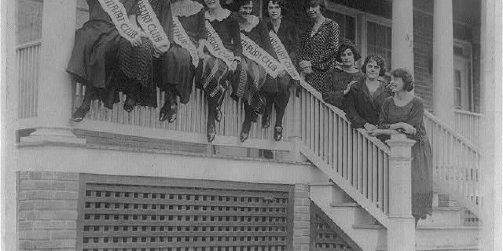A group of women sit on the railing of a porch wearing banners across their chests that read "Anti-flirt club"