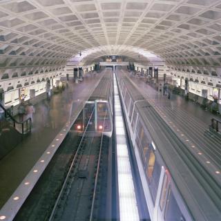 An overhead view of a DC Metro stop with trains running in either direction and blurry people moving around on the platforms. The signature egg carton-esque ceiling arches overhead.