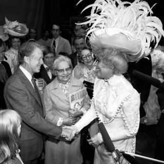 Jimmy Carter shakes actress Carol Channing's hand. They are both grinning widely, Channing is wearing an elaborate white costume, topped with a great hat spewing white feathers in all directions.