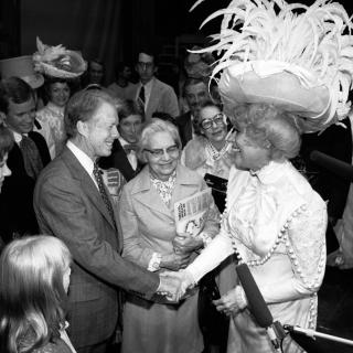 Jimmy Carter shakes actress Carol Channing's hand. They are both grinning widely, Channing is wearing an elaborate white costume, topped with a great hat spewing white feathers in all directions.