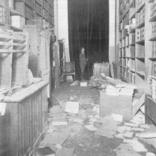 Bookshelves on both sounds of a long hallway are completely disrupted. Books litter the floor, and a man in a suit stands at the end of the hall.