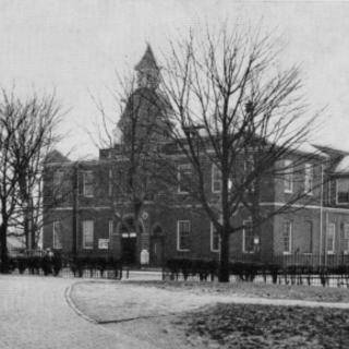 A monochrome photograph featuring the Anne Arundel County courthouse in the background. A path curves in the foreground. The dome is visible behind a screen of tree branches.