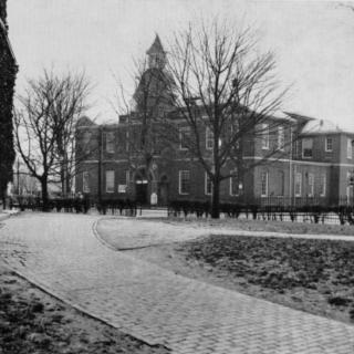 A monochrome photograph featuring the Anne Arundel County courthouse in the background. A path curves in the foreground. The dome is visible behind a screen of tree branches.