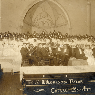 The Samuel Coleridge-Taylor Choral Society, a chorus of around 200 people, posing with orchestra and composer Samuel Coleridge-Taylor at a concert in 1906. 