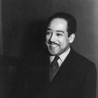 A black-and-white photo of Langston Hughes wearing a suit and smiling against a dark background