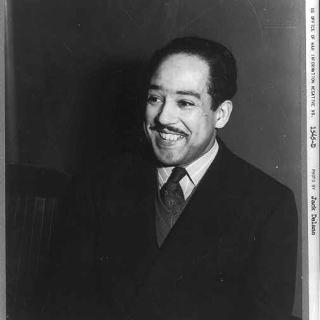 A black-and-white photo of Langston Hughes wearing a suit and smiling against a dark background