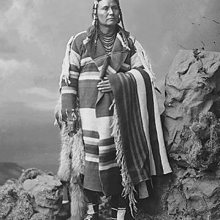 Black and white portrait photo of 39-year-old Chief Joseph standing in front of rock backdrop, wearing striped blanket.