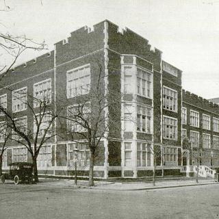 Streetview image of Dunbar High School circa 1917. Castle-like red brick and stone trimmed structure with cars parked on left side.