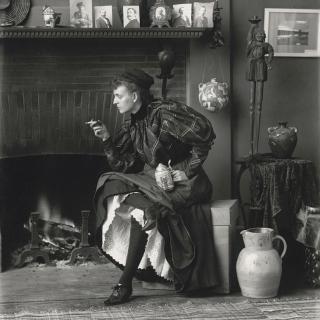 Full body photograph of Frances Benjamin Johnston seated, wearing a dark formal dress and holding a cigarette, 1896. (Source: Library of Congress)