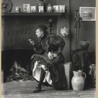 Full body photograph of Frances Benjamin Johnston seated, wearing a dark formal dress and holding a cigarette, 1896. (Source: Library of Congress)