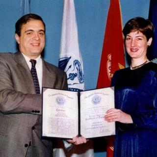 Man and woman posing and smiling for camera in front of flags while woman holds open certificate.