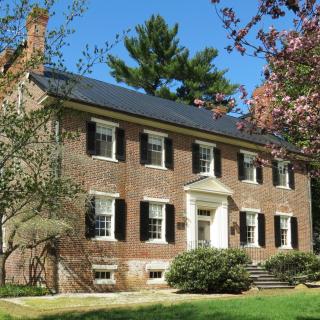 The picture is of a 2 story brick house with a black roof and 4 chimneys, with two on either side. The front of the house is slightly obscured by large bushes that sit along the front and side, and trees which are scattered around the house