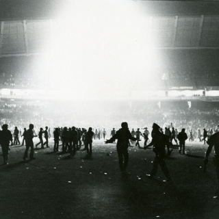 Light shines down from the roof of the Senators baseball stadium, illuminating hundreds of fans storming the field.