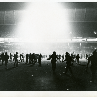 Light shines down from the roof of the Senators baseball stadium, illuminating hundreds of fans storming the field.