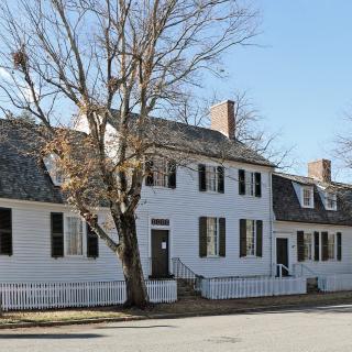 A photo of a large white house with black roof and shutters. The center of the house has a second floor and on either side of it dormer windows are unevenly placed along the roof