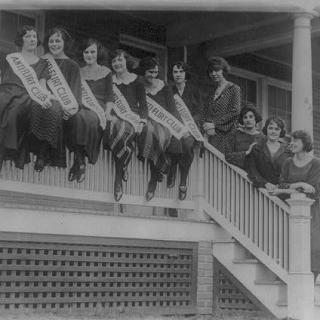 A group of women sit on the railing of a porch wearing banners across their chests that read "Anti-flirt club"