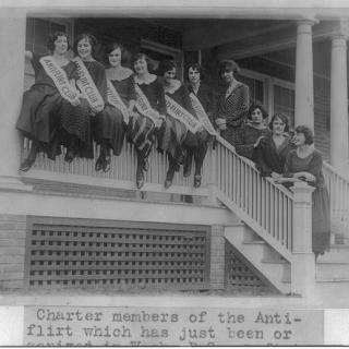 A group of women sit on the railing of a porch wearing banners across their chests that read "Anti-flirt club"