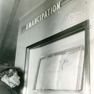 Elderly African American woman look at document in case on the wall. The word Emancipation is displayed over the case.