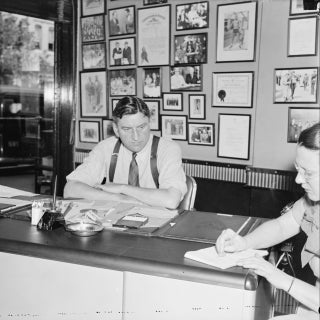 Business man wearing tie sits at a desk talking to a woman at far right of frame, who is taking notes.