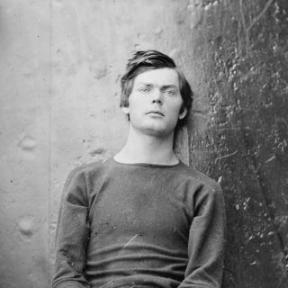Black and white photo of young man sitting against a wall and wearing handcuffs.