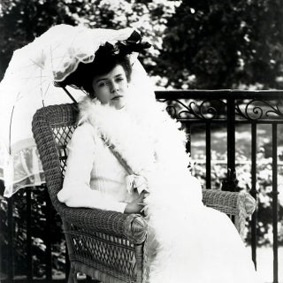 Black and white photo of young woman in white dress and white parasol sitting in chair.
