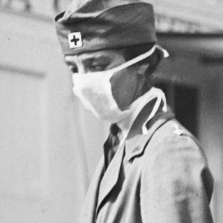This is a black and white photograph of a woman wearing a mask, she wears a hat with the familiar plus sign associated with the Red Cross. Despite the mask covering most of her face, she wears a serious expression. 