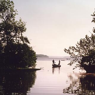 A photograph of the Potomac river. There are two trees on either side of the image. There looks to be two men on a dock in the distance. The water is flat. 