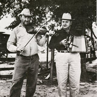 Two men standing in the foreground. The left person is playing a violin while the right person is playing an accordion. 