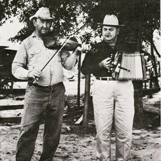 Two men standing in the foreground. The left person is playing a violin while the right person is playing an accordion. 