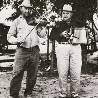 Two men standing in the foreground. The left person is playing a violin while the right person is playing an accordion. 