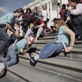 Jennifer Keelan-Chaffins wears a bandana as she crawls up the U.S. Capitol steps on her hands and knees. On her left is a man bending over and on her right a woman moves up the stairs on her back. All three protesters wear matching light blue shirts from the ADAPT organization. In front of them are media personnel with cameras and microphones recording the protest. ​​​​​[Photo Credit: Associated Press]
