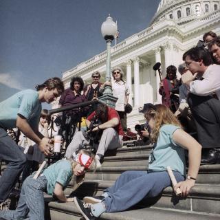 Jennifer Keelan-Chaffins wears a bandana as she crawls up the U.S. Capitol steps on her hands and knees. On her left is a man bending over and on her right a woman moves up the stairs on her back. All three protesters wear matching light blue shirts from the ADAPT organization. In front of them are media personnel with cameras and microphones recording the protest. ​​​​​[Photo Credit: Associated Press]
