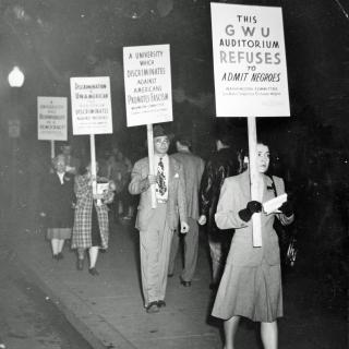 A photograph of people protesting. They are both men and women and they wear clothes from the 1940s. They hold signs that say things such as “a university that discriminates against Americans promotes fascism.”
