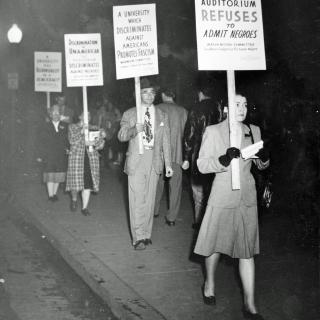 A photograph of people protesting. They are both men and women and they wear clothes from the 1940s. They hold signs that say things such as “a university that discriminates against Americans promotes fascism.”