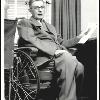 A black-and-white photo. Hugo Deffner, a white man in his 60s, sits in his wheelchair at a desk. He is holding a piece of paper in one hand and wearing a suit. He is looking at the camera.