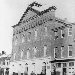 A black-and-white photograph of Fords Theatre in 1868. The building is three stories, brick, with tall windows across every floor. The roof is pointed. There are other buildings attached on either side.