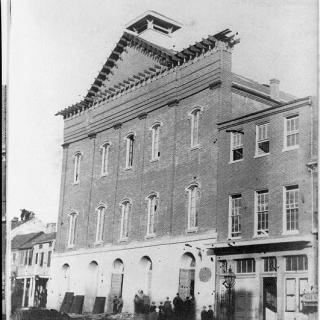 A black-and-white photograph of Fords Theatre in 1868. The building is three stories, brick, with tall windows across every floor. The roof is pointed. There are other buildings attached on either side.
